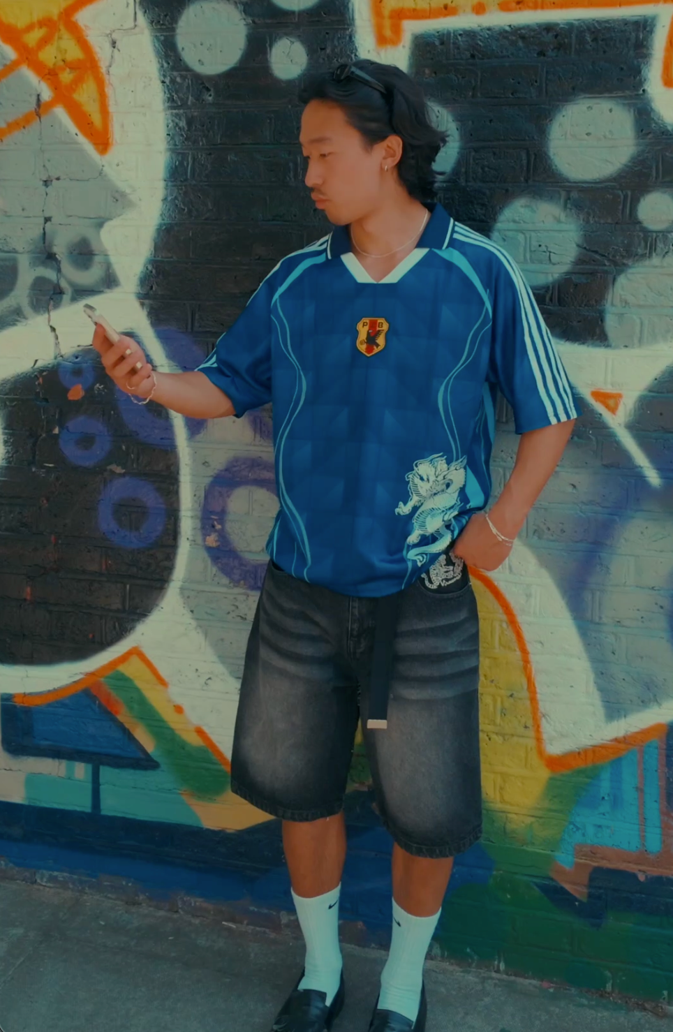 Man wearing navy blue football jersey with white side stripes, PZB shield badge and small graphic on the front, styled with grey denim shorts, white socks and black loafers, standing against a colourful graffiti wall