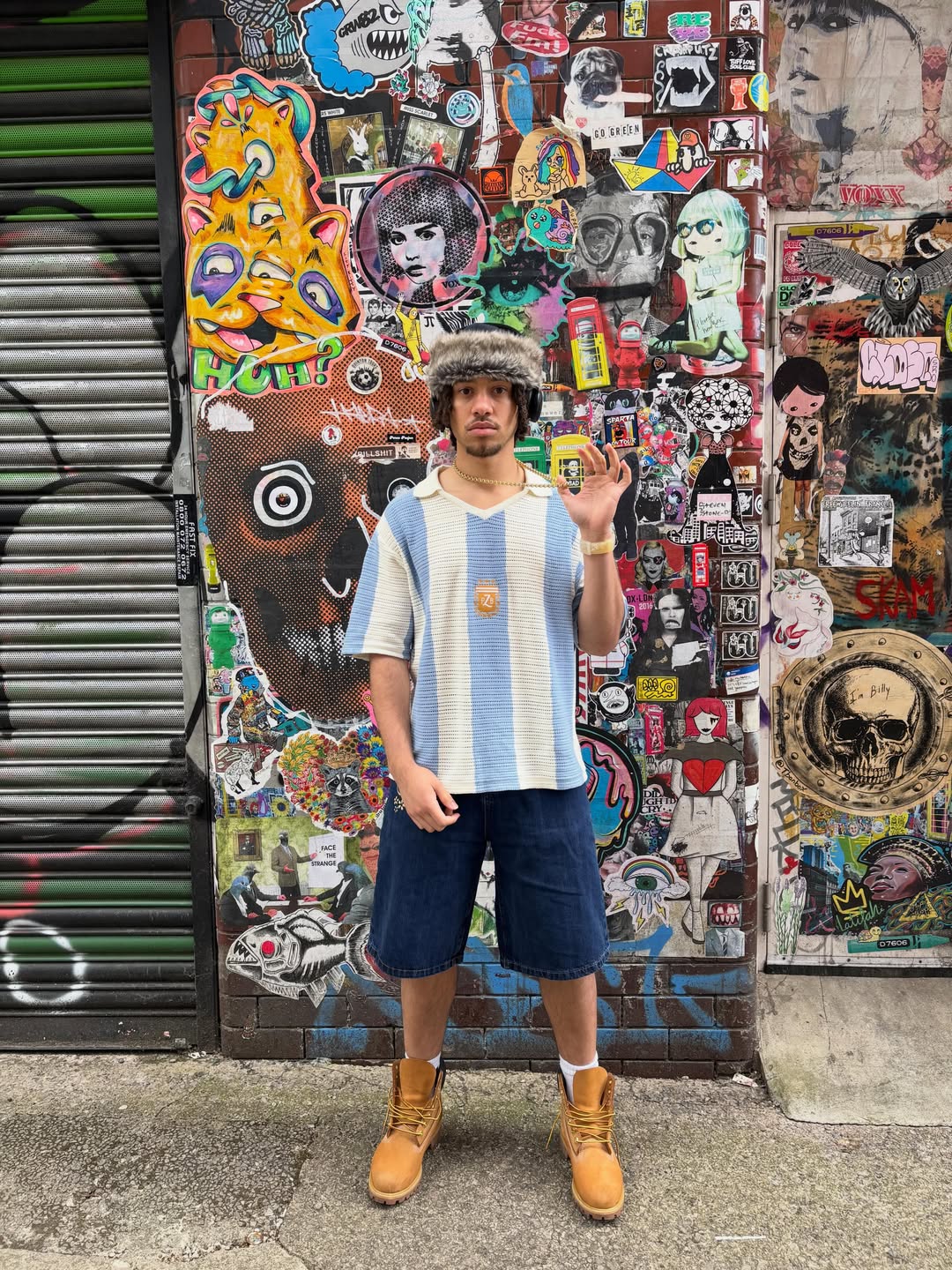 Front view of man wearing light blue and white striped Argentina knit football jersey, styled with denim shorts, white socks, tan Timberland boots and a fur trapper hat, standing against a sticker-covered wall