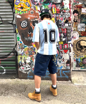 Back view of man wearing light blue and white striped Argentina knit football jersey with black number 10 and Maradona face graphic below the collar, styled with denim shorts, white socks and tan Timberland boots, standing against a sticker-covered wall