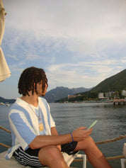 Man wearing light blue and white striped Argentina knit football jersey with PZB badge, styled with black striped shorts and chain necklace, seated by a waterfront with mountains and a harbour in the background