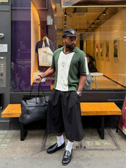 Man wearing an olive green knit polo layered over a white graphic tee, styled with dark wide-leg shorts, white socks, black loafers and a black cap, holding a black leather bag outside a café on a city street