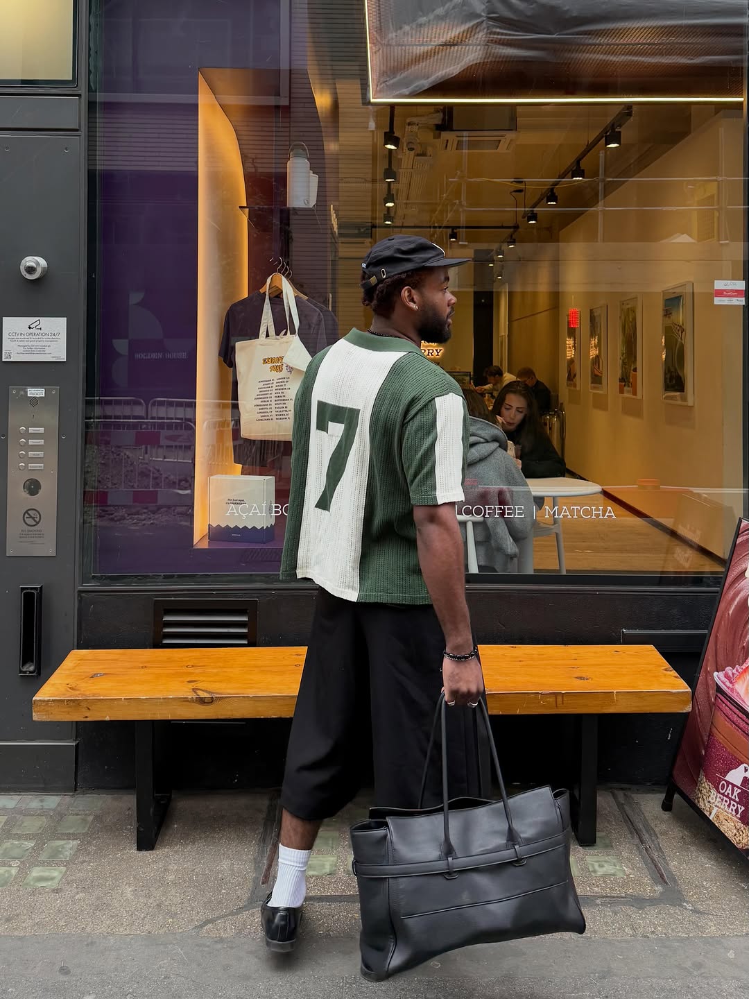 Back view of man wearing green and white colour-block football jersey with number 7, styled with dark wide-leg shorts, white socks, black loafers and a black cap, carrying a black leather tote bag outside a city café