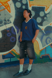 Man wearing navy blue football jersey with PZB shield badge and white V-neck collar, styled with grey denim shorts, white socks and black loafers, standing against a colourful graffiti mural