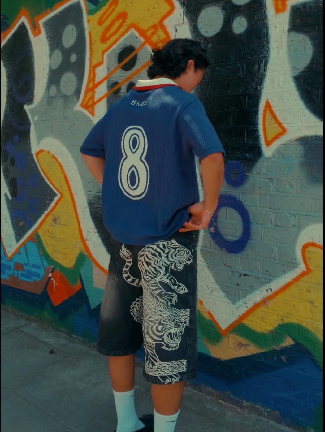 Back view of man wearing navy blue football jersey with white number 8, styled with dark tiger-print shorts and white socks, standing against a colourful graffiti mural