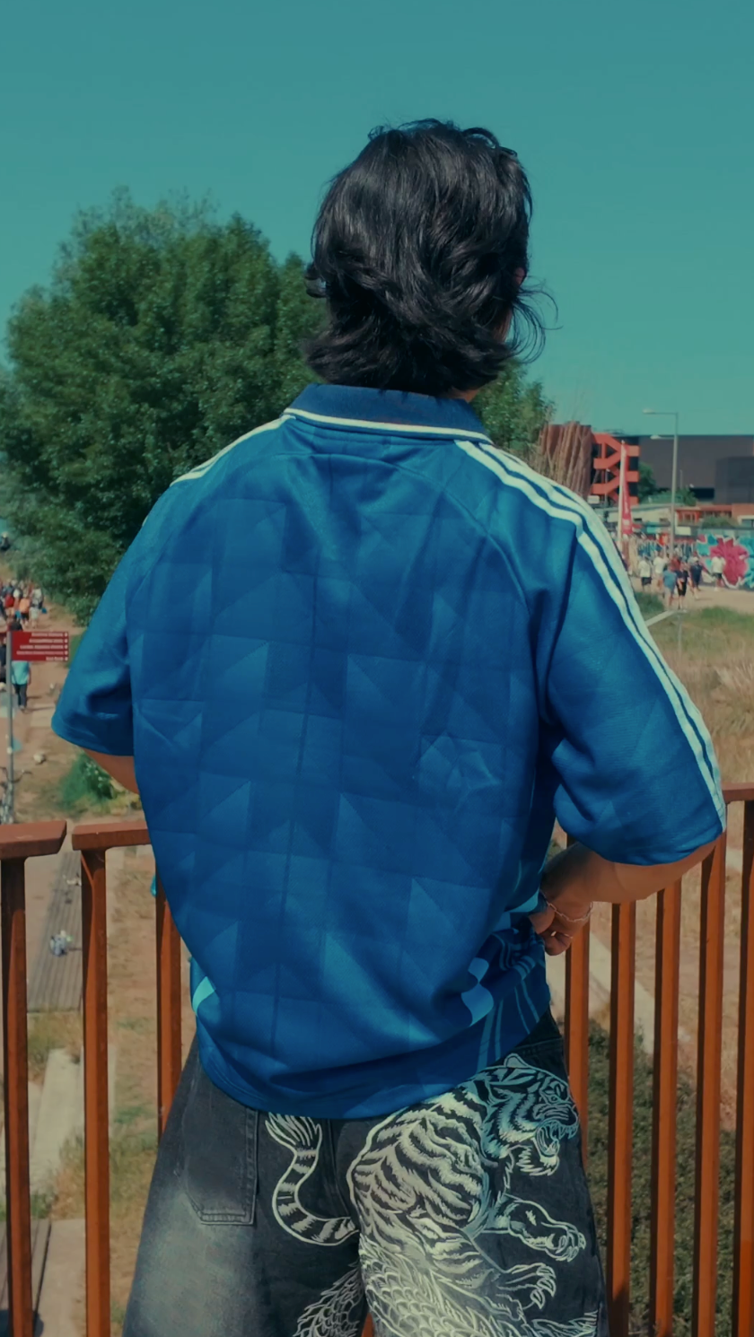 Back view of man wearing navy blue football jersey with white side stripes, styled with tiger-print shorts, leaning against a wooden railing with an outdoor venue in the background