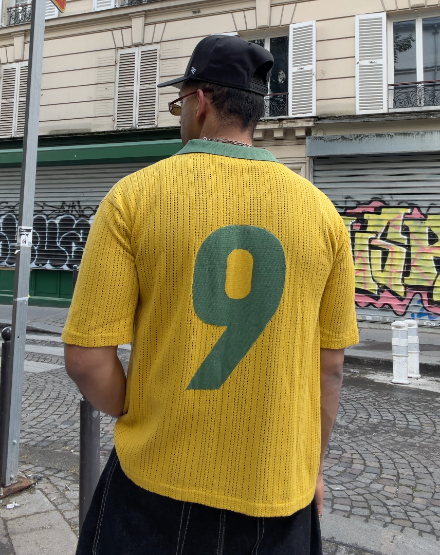 Back view of man wearing yellow knit football jersey with green number 9 and green collar, styled with black wide-leg trousers and a black cap, on a cobblestone street in Paris