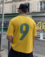 Back view of man wearing yellow knit football jersey with green number 9 and green collar, styled with black wide-leg trousers and a black cap, on a cobblestone street in Paris