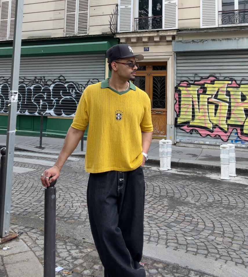 Man wearing yellow knit football jersey with green collar and PZB badge, styled with black wide-leg trousers and a black cap, standing on a cobblestone street in Paris