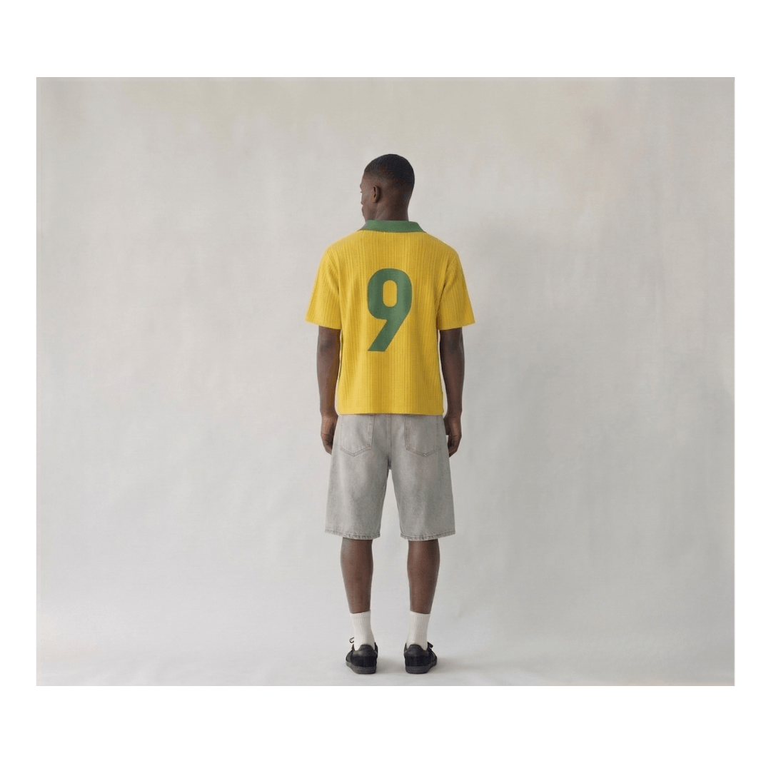 Rear view of a man wearing a Brazil 1998 retro-style yellow knitwear shirt. The shirt features a large, dark green number "9" on the back and a matching green ribbed collar. The fabric has a distinct vertical ribbed knit texture, styled with light grey cargo shorts and black sneakers on a plain studio background.