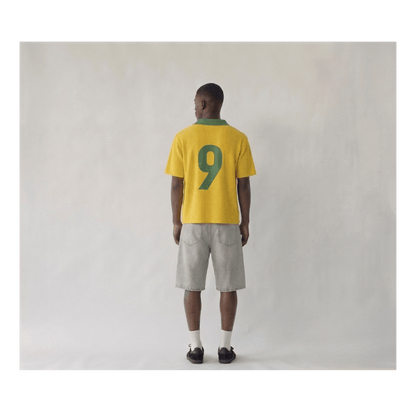 Rear view of a man wearing a Brazil 1998 retro-style yellow knitwear shirt. The shirt features a large, dark green number "9" on the back and a matching green ribbed collar. The fabric has a distinct vertical ribbed knit texture, styled with light grey cargo shorts and black sneakers on a plain studio background.