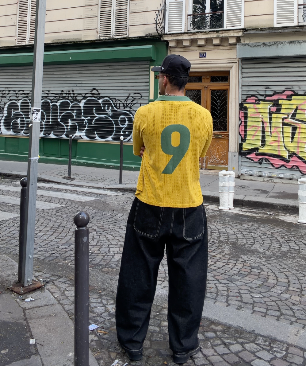 Back view of model wearing a ParkzBus yellow Brazil-inspired waffle-knit polo with large green number 9, black wide-leg trousers with contrast stitching, stood on a Parisian cobblestone street with graffiti shutters