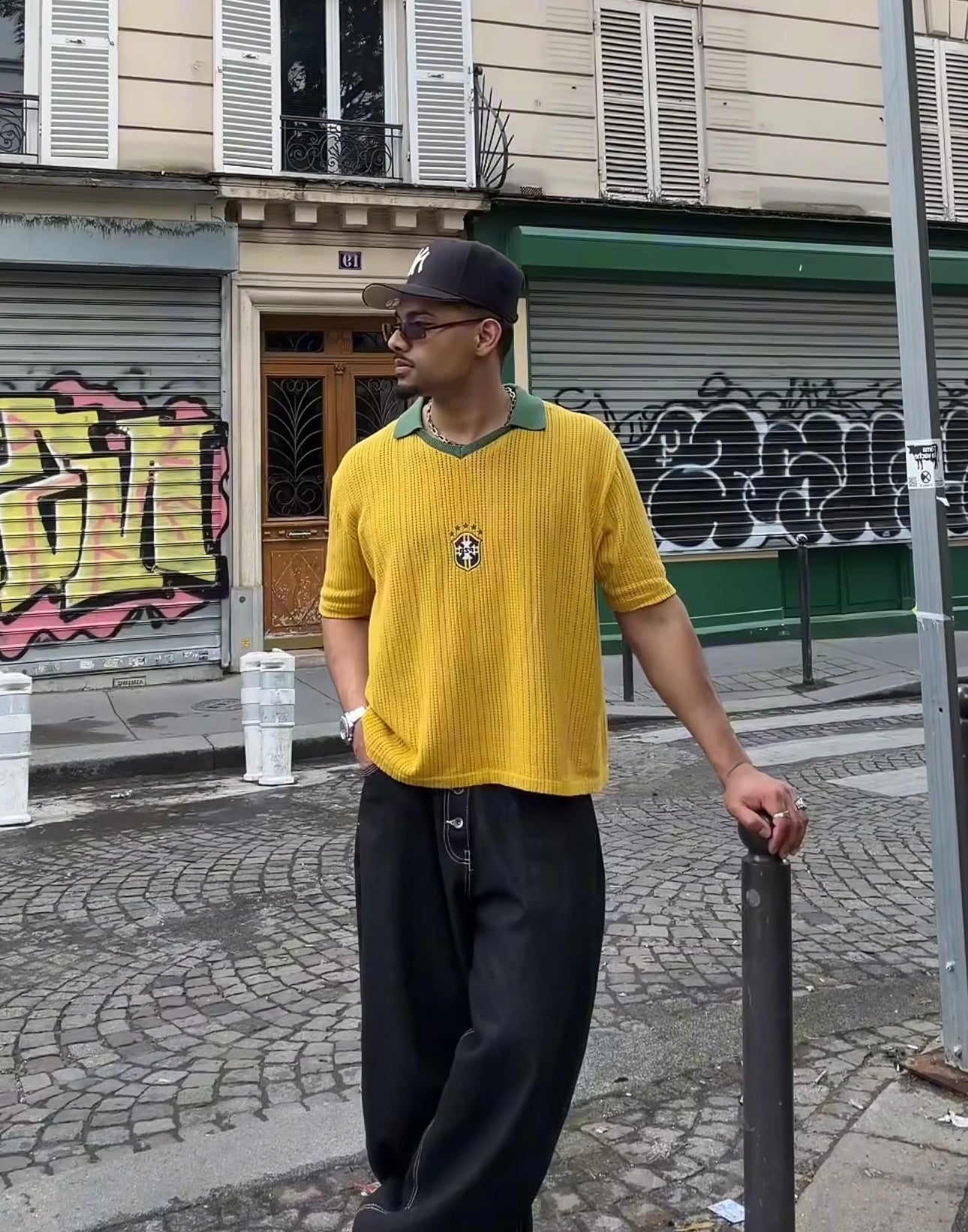 Model wearing a yellow Brazil-inspired ParkzBus knit polo with green collar, black wide-leg trousers and a New Era cap, stood on a Parisian cobblestone street with graffiti shutters.