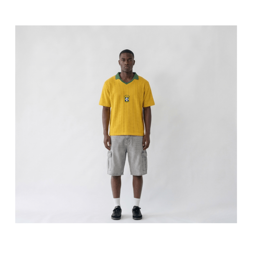 Front view of a man wearing a Brazil retro-style yellow knitwear football shirt. The shirt features a green ribbed V-neck collar and the official CBF badge centred on the chest. The fabric has a distinct vertical ribbed knit texture, styled with light grey cargo shorts and black sneakers on a plain white studio background.