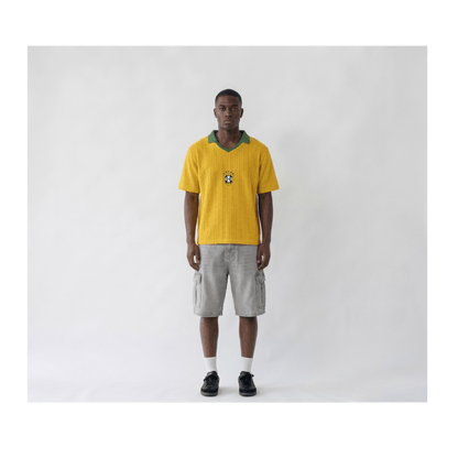 Front view of a man wearing a Brazil retro-style yellow knitwear football shirt. The shirt features a green ribbed V-neck collar and the official CBF badge centred on the chest. The fabric has a distinct vertical ribbed knit texture, styled with light grey cargo shorts and black sneakers on a plain white studio background.